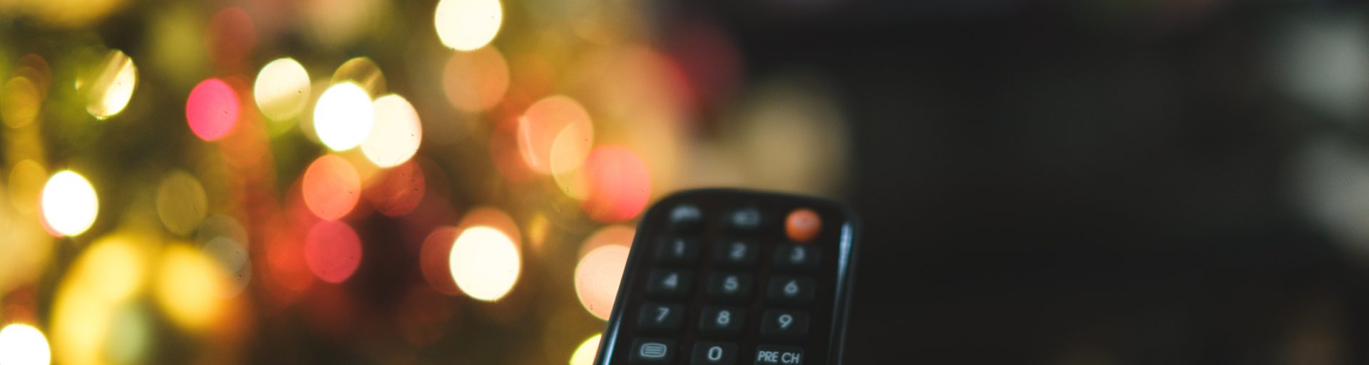 Subjective close-up of a hand holding the remote control, with a background of led lights of the Christmas tree and TV on. I choose with the remote control what to watch on television during the holidays